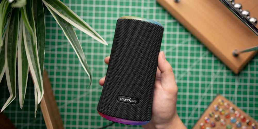 اسپیکر بلوتوثی انکر مدل Soundcore Flare 2 با یک دست روی یک سطح صاف با یک گیاه و اشیاء چوبی در پسزمینه نگه داشته میشود.
