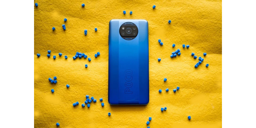 موبایل پوکو ایکس 3 NFC آبی روی پارچه ای زرد رنگ که مهره هایی آبی رنگ در اطراف آن ریخته شده است