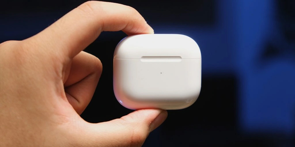 یک دست قاب شارژ Apple AirPods (نسل سوم) را می‌گیرد تا نمای بیرونی سفید ساده و فرورفتگی مینیمالیستی آن را نشان دهد