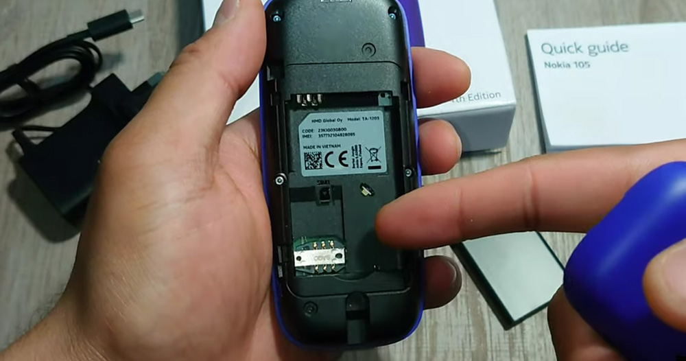 گوشی نوکیا 105 جدید در دست قرار دارد. درب پشت آن و باتری آن روی دستگاه قرار ندارد و دست دیگر در حال اشاره به جایگاه سیم کارت می باشد