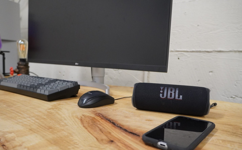 یک بلندگوی بلوتوث JBL Flip 6 که روی یک میز چوبی در کنار گوشی هوشمند در سمت راست و یک مانیتور، صفحه کلید و ماوس در سمت چپ آن قرار دارد.